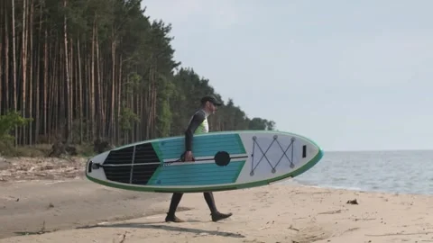Man walking down beach to water with stand up paddle board in his hands. SUP Stock Footage 244605629