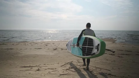 Man walking down beach to water with stand up paddle board in his hands. SUP Stock Footage 244606238