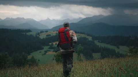 Man walking on grass in meadow hill overlooking mountain peaks and rainy clouds Stock Footage 282444860