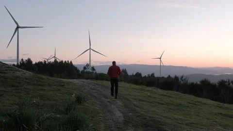 Man walking on a hill towards wind turbines at sunset Stock Footage 344531141