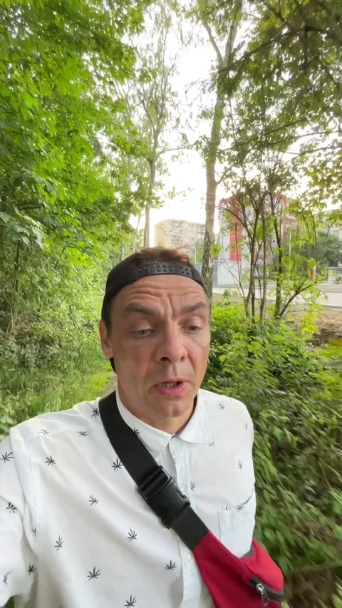 Man walking leafy urban path, selfie-style portrait outdoors, red crossbody bag Vídeos de archivo 344586332