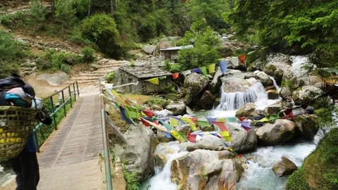 Man walking over water stream on a bridge with Tibetan prayer flags waving Stock Footage 77445988