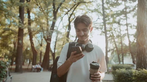 Man walking in park with coffee and phone, wearing backpack and headphones 库存影片 339216566