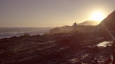 Man walking on rocks by the ocean as the sun is setting Stock Footage 279797593