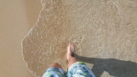 Man walking on sandy beach, waves gently touching his feet in a peaceful Stock Footage 287445843