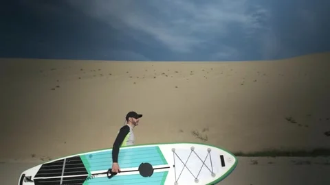 Man walking on shore with stand up paddle board along big dunes in desert. SUP Stock Footage 244605253