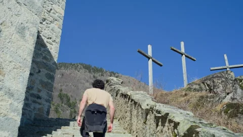 Man Walking Up Stone Stairs Behind Church Towards Crosses in Mountains Stock Footage 316420498