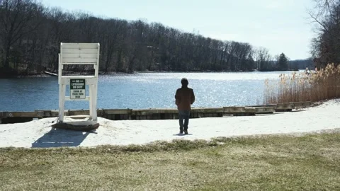 Man Walking Towards Sparkling Lake Shore with Lifeguard Chair - 4K Stock Footage 331746423