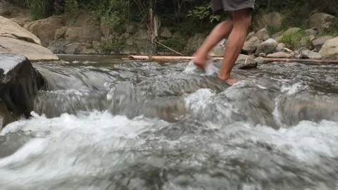 A man walks across a river or wild stream barefoot. Steady shot of a clear,.. Video stock 334486602