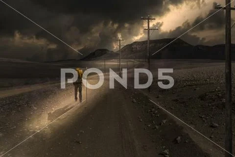A man walks along an empty road in a sunbeam shining through dark clouds, compos Stock Photos
