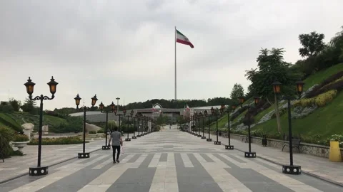 Man walks along lamppost-lined walkway toward building beneath Iranian flag Stock Footage 335935712
