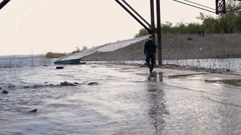 A man walks along the riverbank.In the background is a rocky shore,boat,forest. Stock Footage 130727853