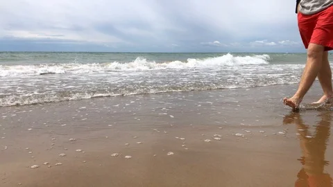 Man walks along sandy beach with sea waves, sailboard far away Stock Footage 93548484