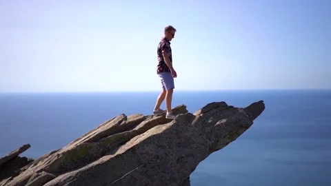 A man walks along a sheer cliff jutting out from the sea against the blue sky Stock Footage