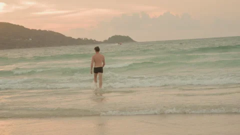 Man walks into evening waves. Man with determined stride faces ocean at sunset Stock Footage 332931674