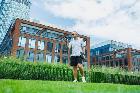 Man walks on grass while holding a smartphone near modern buildings during .. Stock Photos