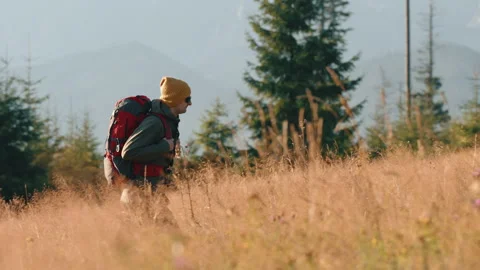 Man walks meadow overlooking mountain peaks lit with sunrise light, red backpack Stock Footage 282446927