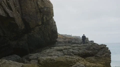 A man walks on rocks on a cliff by the ocean on a cloudy day, in France Stock Footage 229663313