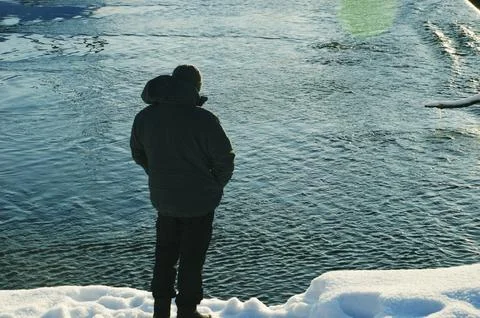 A man in a warm winter gray jacket and a knitted hat stands on the bank of a Stock Photos
