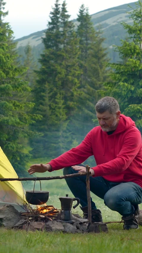 Man warming hands by campfire cooking outdoors in mountain campsite Stock Footage 335438690