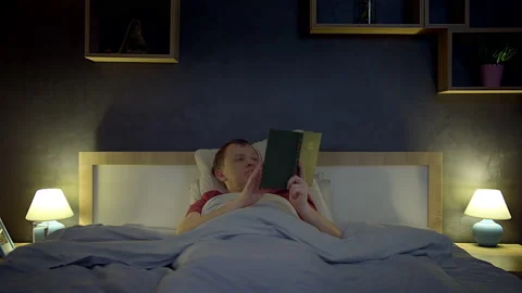 A man was reading a book before going to bed lying in his bed, then took a Stock Footage 209689593