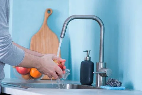 A man washes fruit in a sink in a kitchen with blue walls. Against the backgr Stock Photos