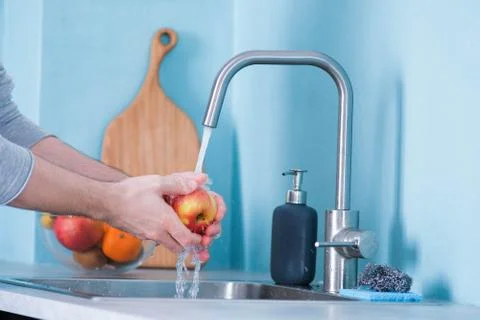 A man washes fruit in a sink in a kitchen with blue walls. Against the backgr Stock Photos