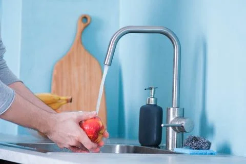 A man washes fruit in a sink in a kitchen with blue walls. Against the backgr Stock Photos