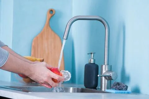 A man washes fruit in a sink in a kitchen with blue walls. Against the backgr Stock Photos