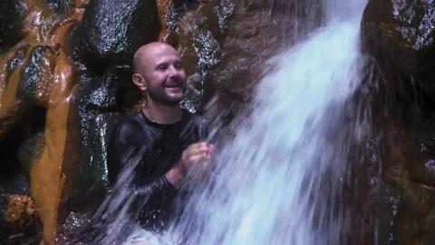 A man washes his face in a waterfall. A large stream of water falls on the man. Vídeo Stock 133460743