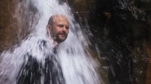 A man washes his face in a waterfall. A large stream of water falls on the man. Stock Footage 133566948