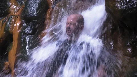 A man washes his face in a waterfall. A large stream of water falls on the man. Vídeos de archivo 133579334