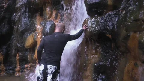 A man washes his face in a waterfall. A large stream of water falls on the man. Vídeos de archivo 133579489