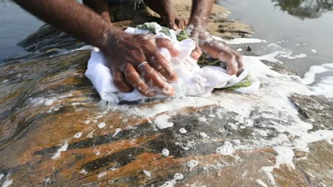 A man is washing clothes by hand on a stone at the riverbank. Видео 333025563