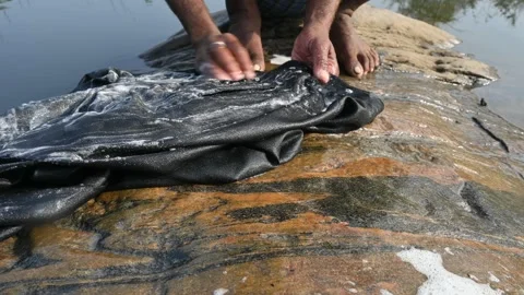 A man is washing clothes by hand on a stone at the riverbank. Video stock 333025602