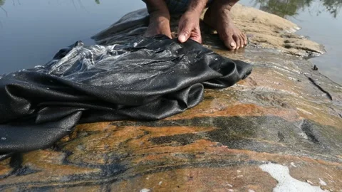 A man is washing clothes by hand on a stone at the riverbank. Video stock 333025970