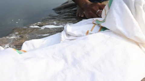 A man is washing clothes by hand on a stone at the riverbank. Vídeos de archivo 333025982