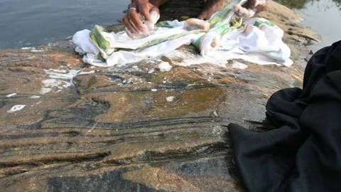 A man is washing clothes by hand on a stone at the riverbank. Vídeos de archivo 333026085