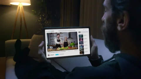 Man watches a cooking tutorial on a tablet while laying on sofa in the livi.. Vídeos de archivo 279595818