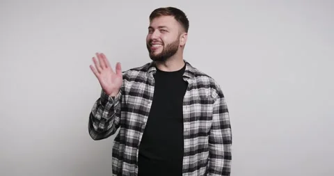 Man waves hello in a simple setting with a plain background and casual clot.. Vídeo Stock 326798233
