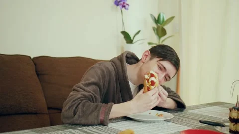 Man wearing bathrobe sitting at table eating hot dog. Male in cozy home Stock Footage 306477577