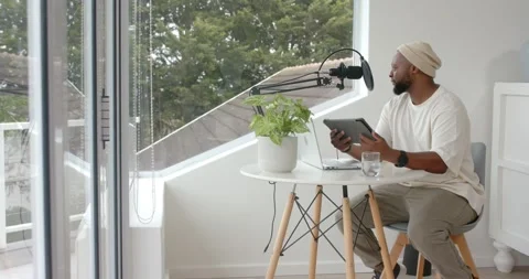 Man wearing beanie sitting at table by window, consulting tablet and speaking Stock Footage 341241735