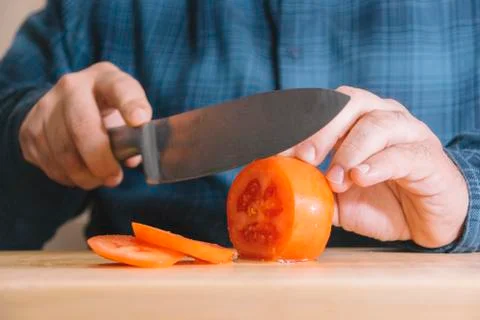 Man wearing a blue shirt slicing a tomato on a wooden board. Cooking concept Stock Photos