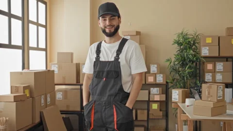 Man wearing cap and overalls hands in pockets next to stacked parcels and b.. Stock Footage 337371862