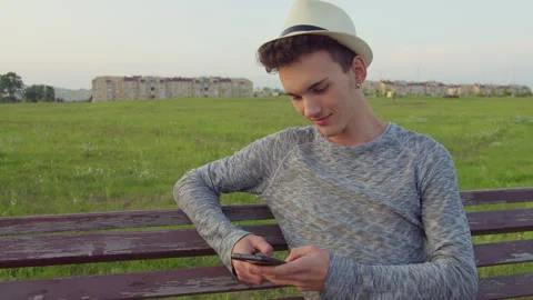 A man wearing a hat sits on a bench with a mobile phone in hand, texting and Stock Footage 312888896