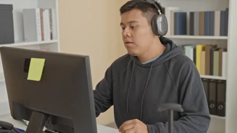 Man wearing headphones types at computer while holding a pen and leaning to.. Stock Footage 347803619