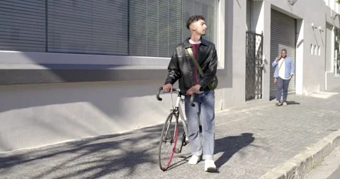 Man wearing leather jacket pushing bicycle with red wheel on sidewalk, pausing Stock Footage 342926799