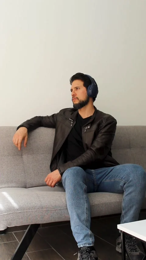 Man wearing a leather jacket sitting on a grey couch listening music Video stock 340583471