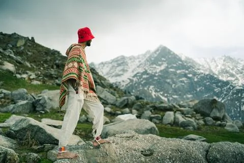 A man wearing a patterned poncho and red hat stands on a rocky mountain slope Stock Photos