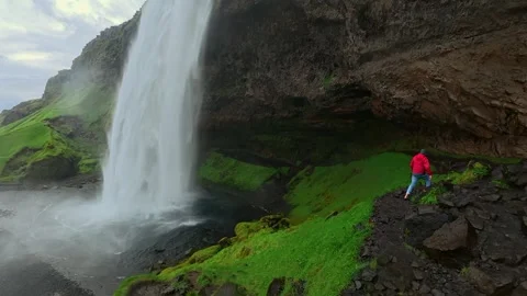 Man wearing red jacket walking on a path behind the Seljalandsfoss waterfall Stock Footage 337949613